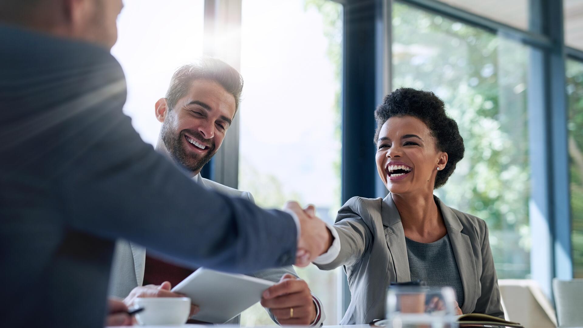 three business people around a table. Two people, a man and a woman, are shaking hands